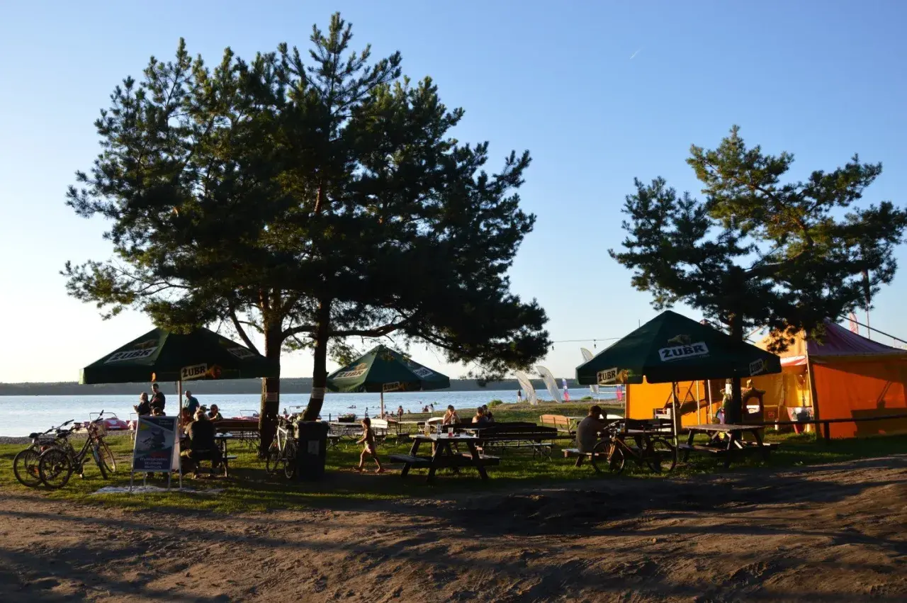 Relaks na plaży Karolinów. Ludzie siedzą pod parasolami, rowery czekają na plaży, a w tle jezioro i namioty.