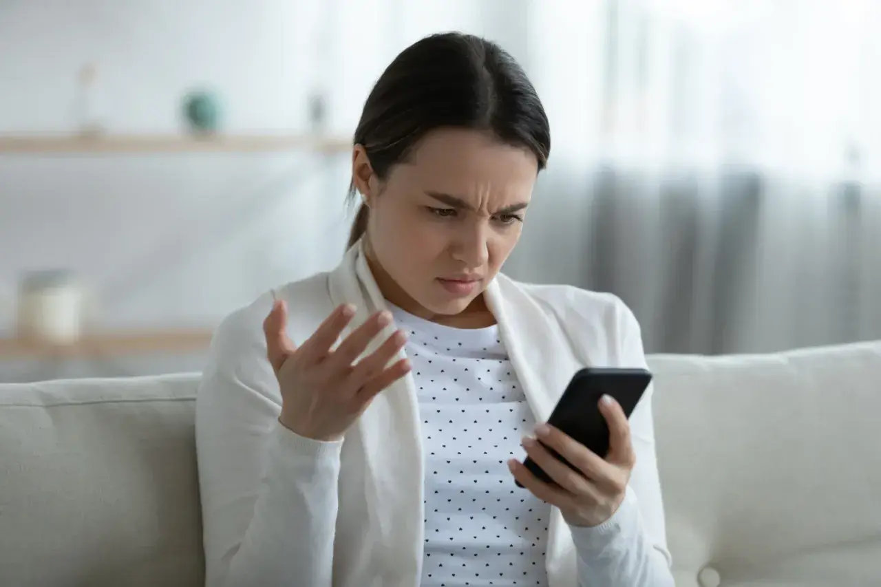Kobieta z irytacją patrzy na telefon, gestykulując ręką. Ewidentnie przeszkadza jej słaby internet w telefonie, uniemożliwiający normalne korzystanie.