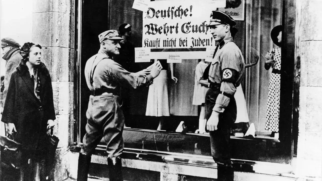 Ein SA-Mann klebt ein Plakat "Deutsche! Wehrt Euch! Kauft nicht bei Juden!" an ein Schaufenster. Dies ist ein Beispiel für den **Alltag im Nationalsozialismus**.