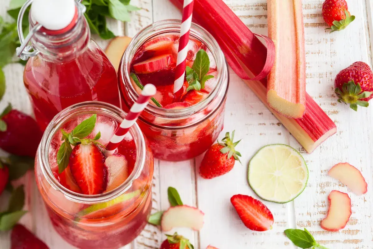 Orzeźwiający sok z rabarbaru z truskawkami i miętą, podany w słoikach z lodem i słomkami. Obok leżą świeże rabarbar, truskawki i plasterki limonki.
