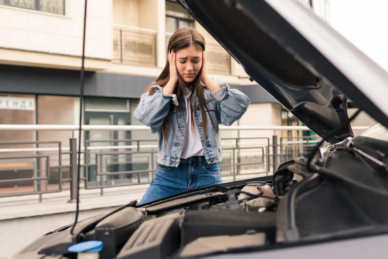 Zrozpaczona dziewczyna z rękami na uszach przy otwartej masce samochodu. Jej auto nie odpala, co ją bardzo frustruje.