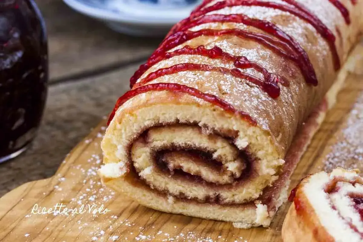Un delizioso rotolo alla marmellata, spolverato di zucchero a velo e decorato con un filo di confettura rossa, pronto per essere gustato.