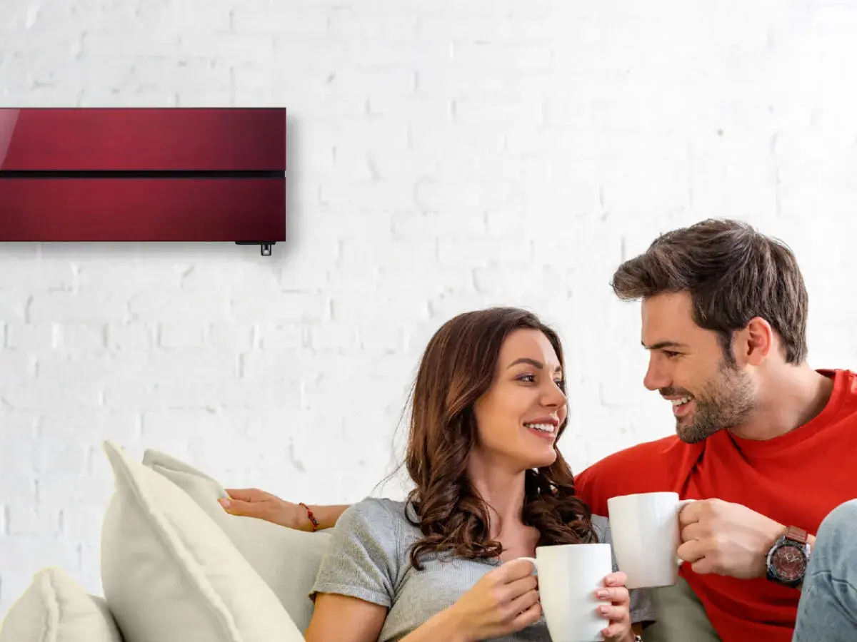 Pareja sonriente en un sofá, disfrutando de café. Arriba, un aire acondicionado rojo moderno garantiza cop eficiencia.