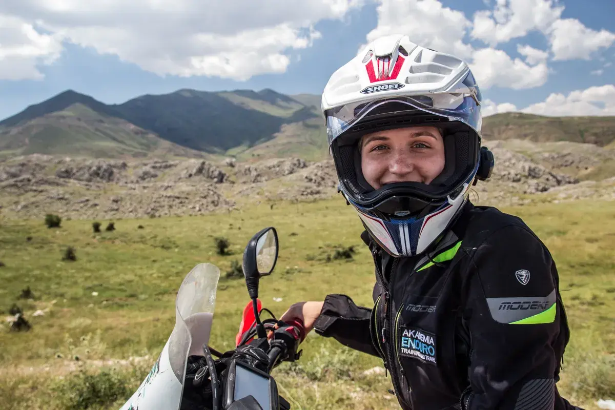 Uśmiechnięta motocyklistka w kasku, gotowa do podróży. W tle górzysty krajobraz, idealny do przygód z **carnet de passage**.