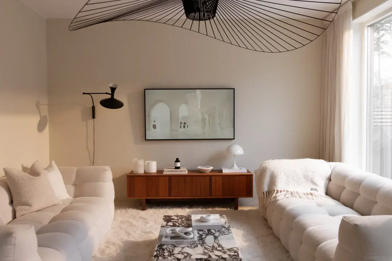 Ett vardagsrum i kall beige ton. Mjuka soffor, en marmorbänk och en träbyrå skapar en lugn atmosfär. En stor taklampa ger ett dramatiskt intryck.