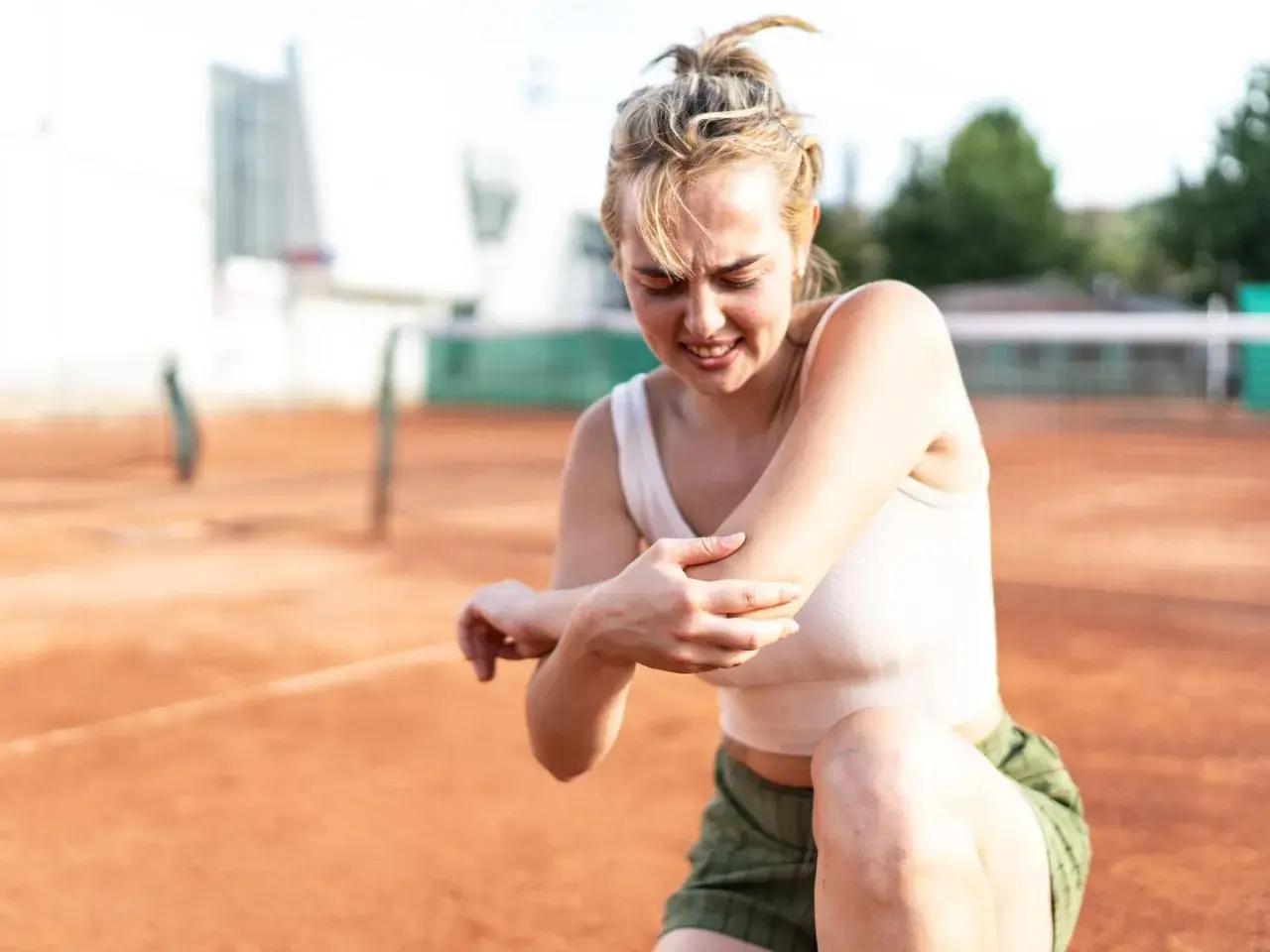 Junge Frau auf einem Tennisplatz hält schmerzgebeugt ihren Ellenbogen. Der Tennisarm wird nicht besser.