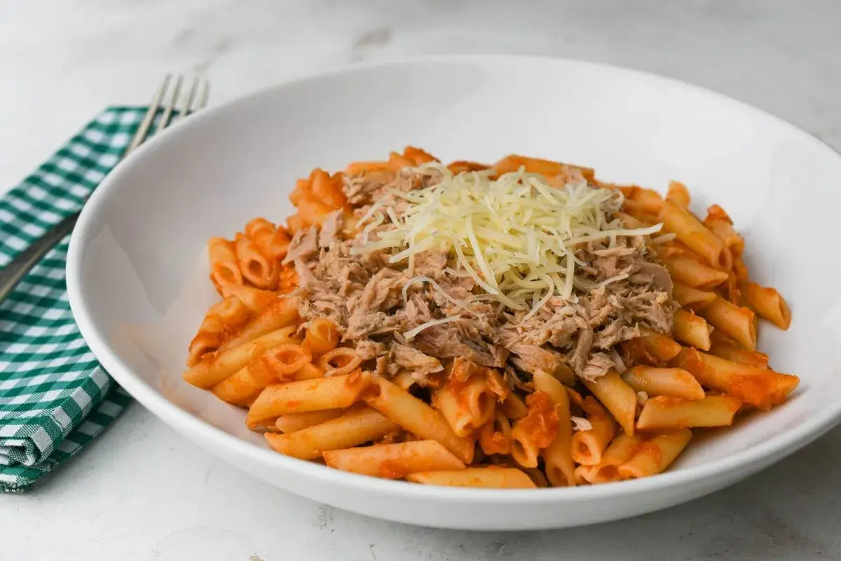 Macarrones con tomate y atún desmenuzado, cubiertos con queso rallado, servidos en un plato blanco.