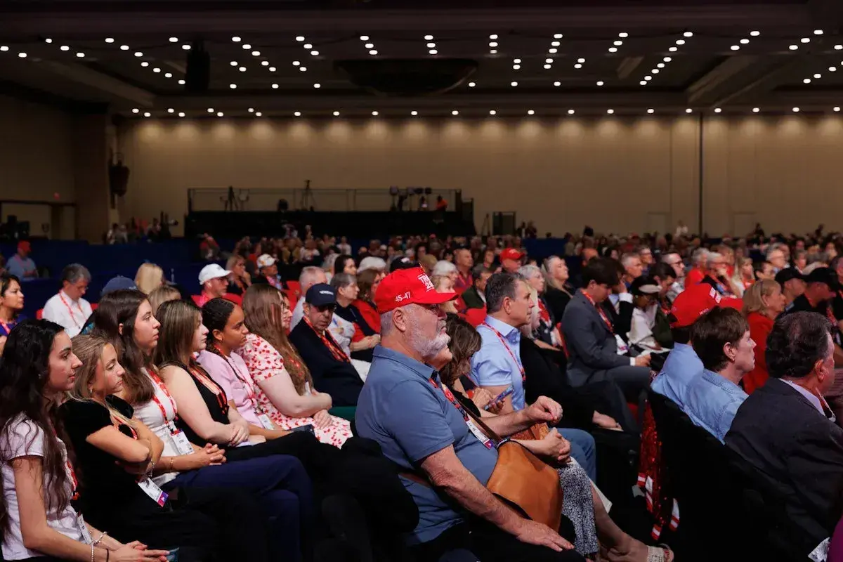 A large audience attends a government conference, with many people wearing red hats.