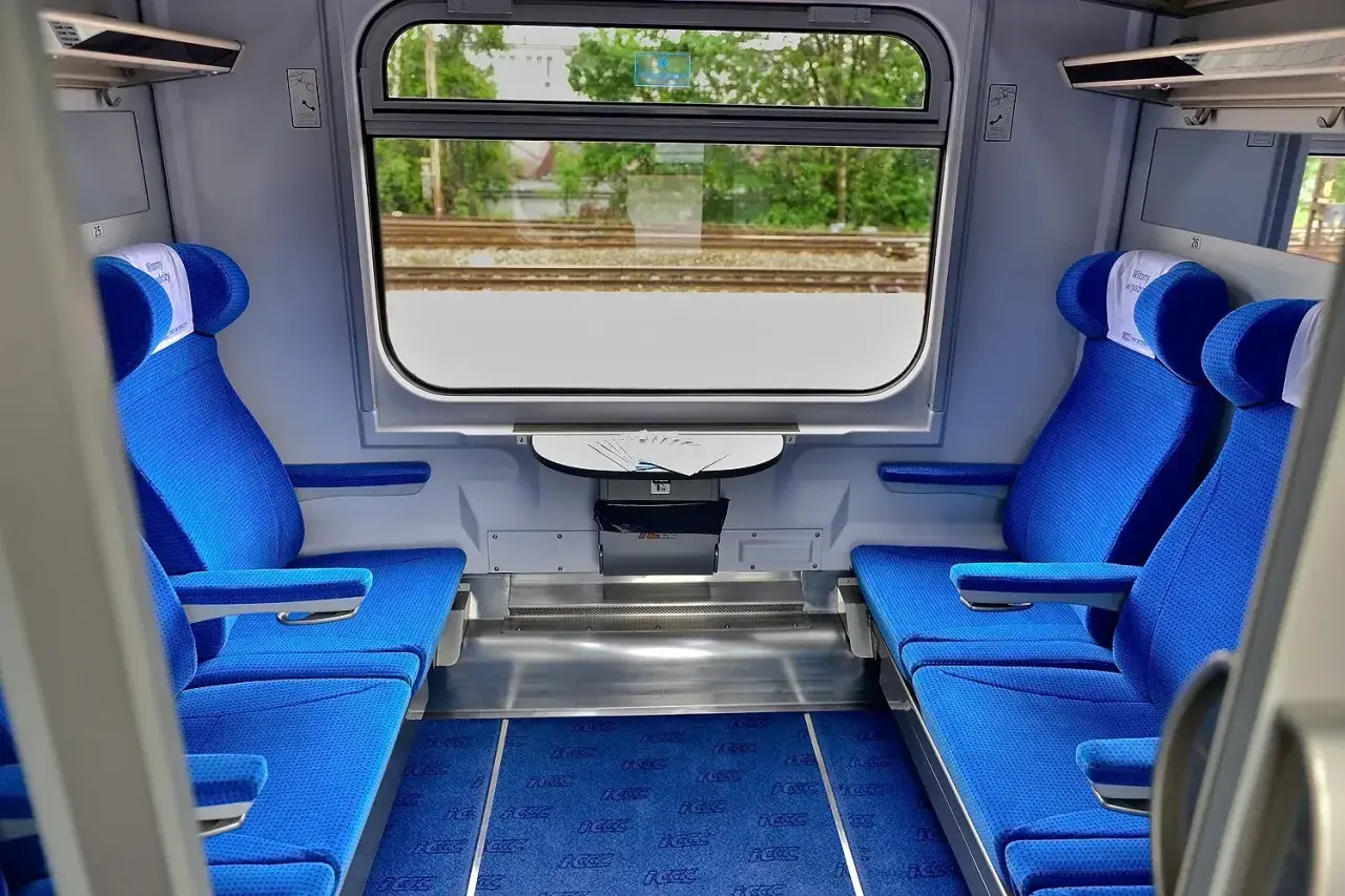 Wnętrze wagonu Intercity z niebieskimi siedzeniami i oknem z widokiem na tory.