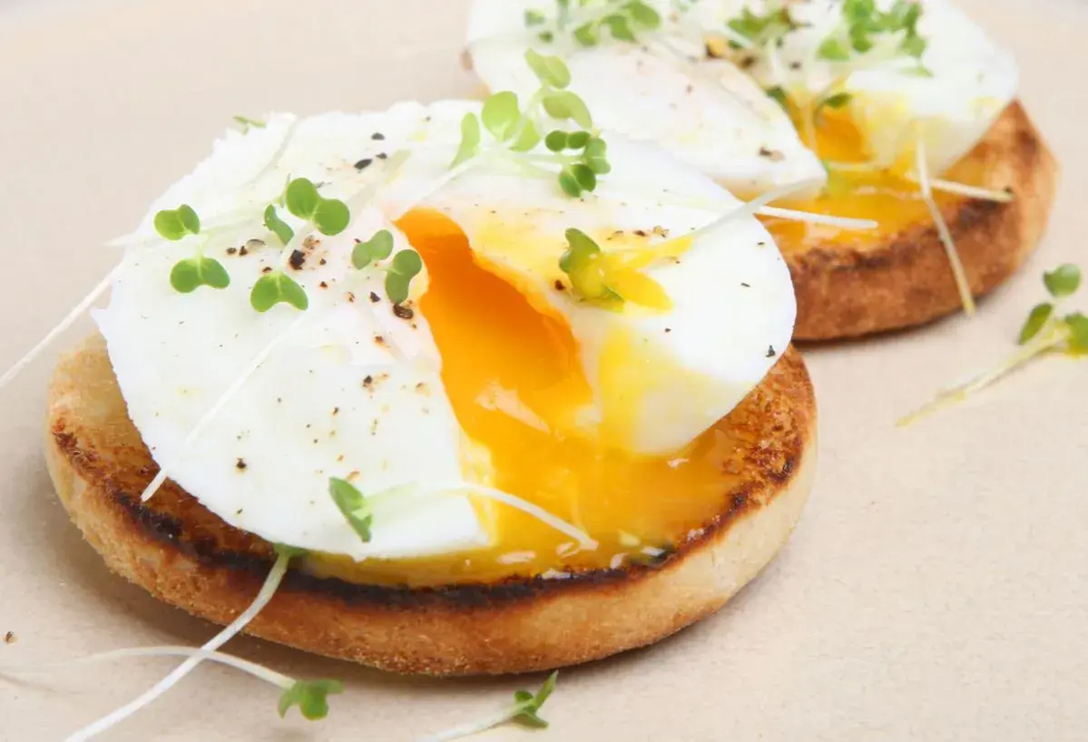 Dos deliciosos huevos poche sobre tostadas inglesas, adornados con brotes verdes y pimienta. La yema dorada fluye, invitando a un bocado perfecto.
