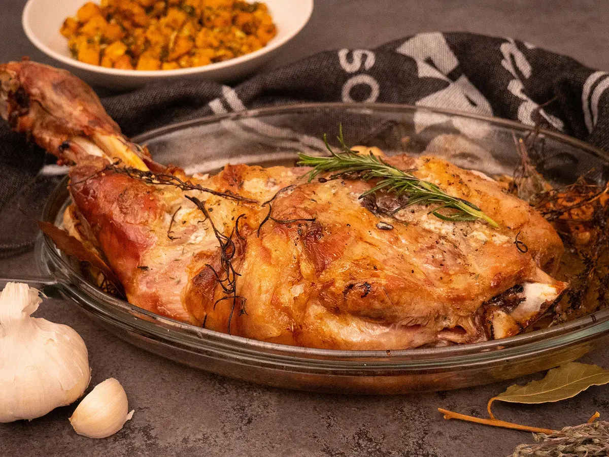 Une succulente épaule d'agneau rôtie, garnie de romarin frais, dans un plat en verre. A côté, de l'ail et une purée de légumes.