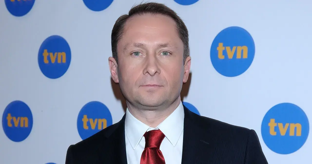 Kamil Durczok w garniturze i czerwonym krawacie na tle logo TVN. Prowadzący Fakty TVN.