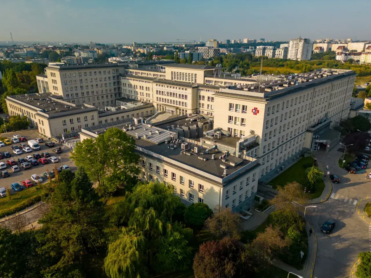 Widok z lotu ptaka na duży kompleks szpitalny z czerwonym krzyżem na fasadzie, otoczony drzewami i parkingiem.