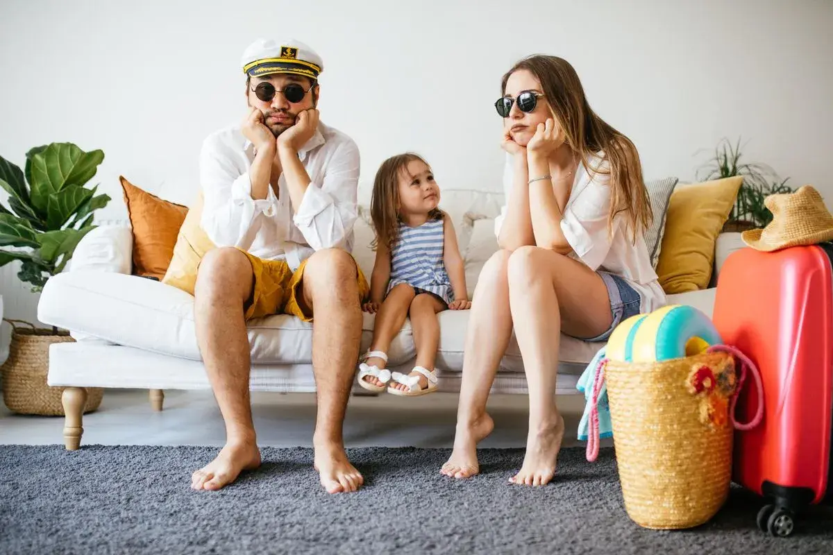 Famille dépitée sur un canapé, valise rouge prête. Annulation location vacances cas de force majeure, les vacances rêvées s'envolent.