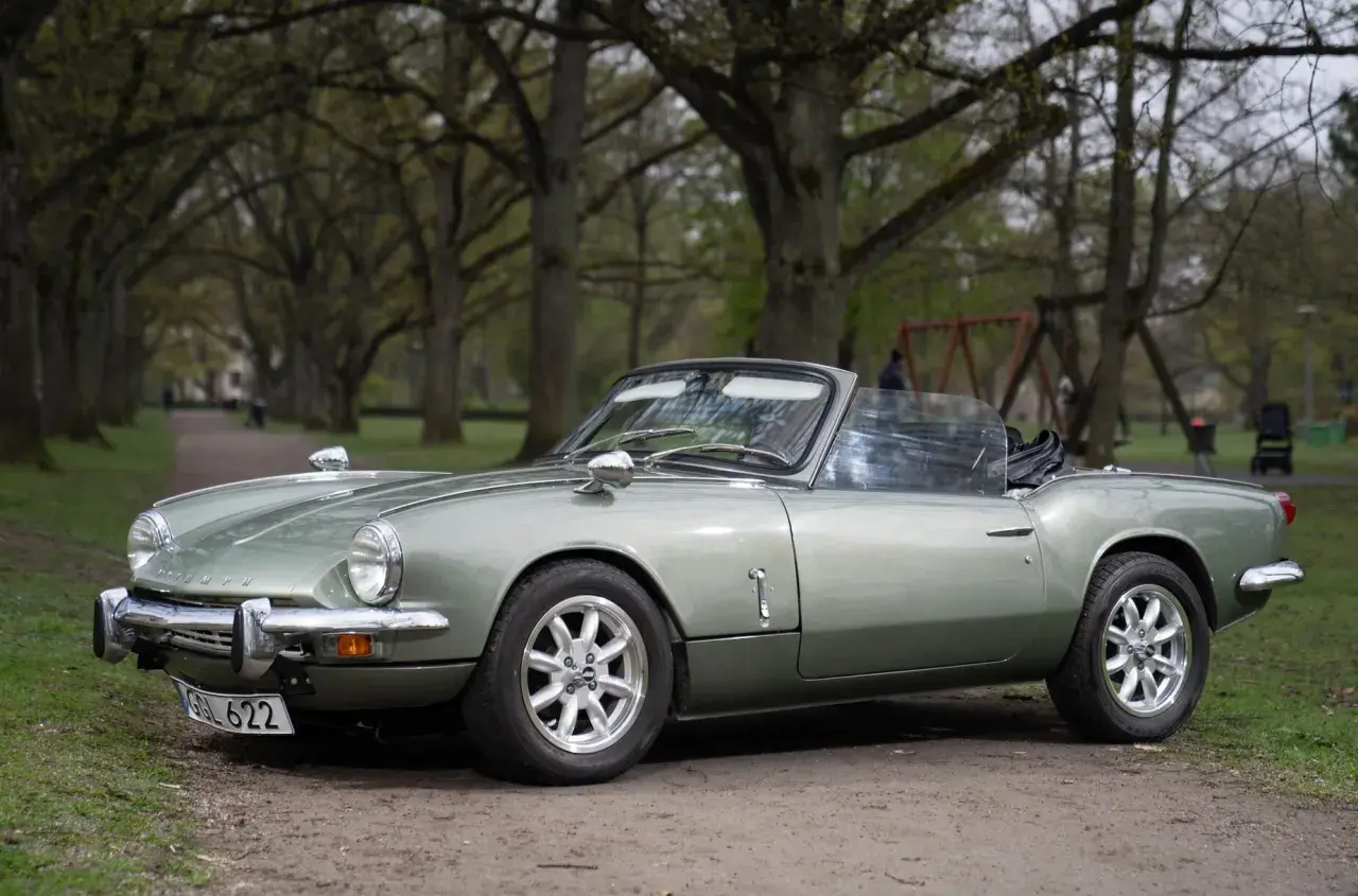 En ljusgrön Triumph Spitfire cabriolet parkerad på en grusväg i en allé.