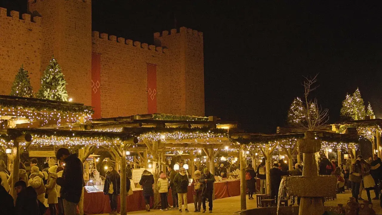 Mercado navideño bajo un castillo iluminado. Gente pasea entre puestos decorados con luces, mostrando como se celebra la navidad.