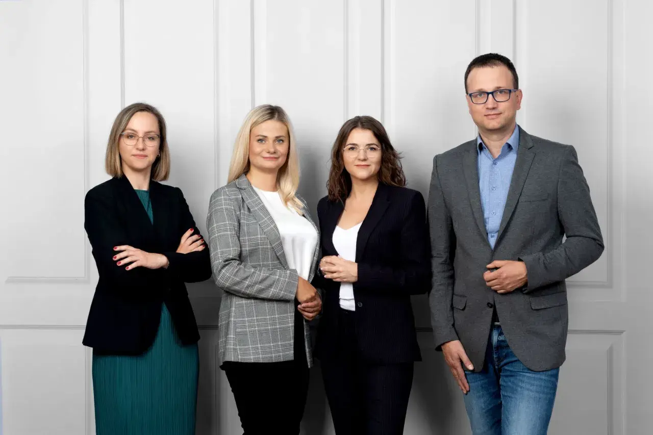 Cztery osoby w strojach biznesowych stoją przed jasną ścianą.