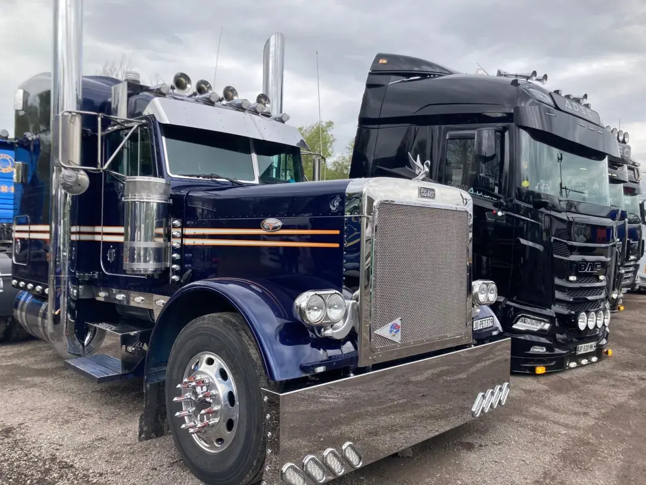 Deux camions américains, un bleu et un noir, brillent sous un ciel couvert. Le bleu, un Peterbilt, arbore des touches orange et beaucoup de chrome.