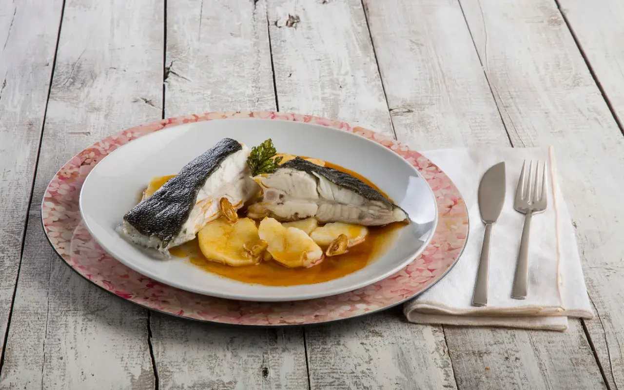Rodaballo gallego servido con patatas y salsa, acompañado de cubiertos y servilleta sobre una mesa de madera.