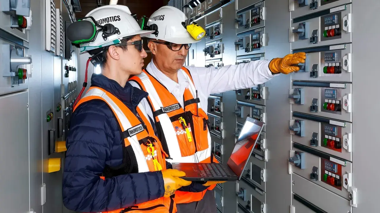Two engineers in a control room, one pointing at a panel, the other holding a laptop, showcasing the characteristics of innovation in electrical systems.