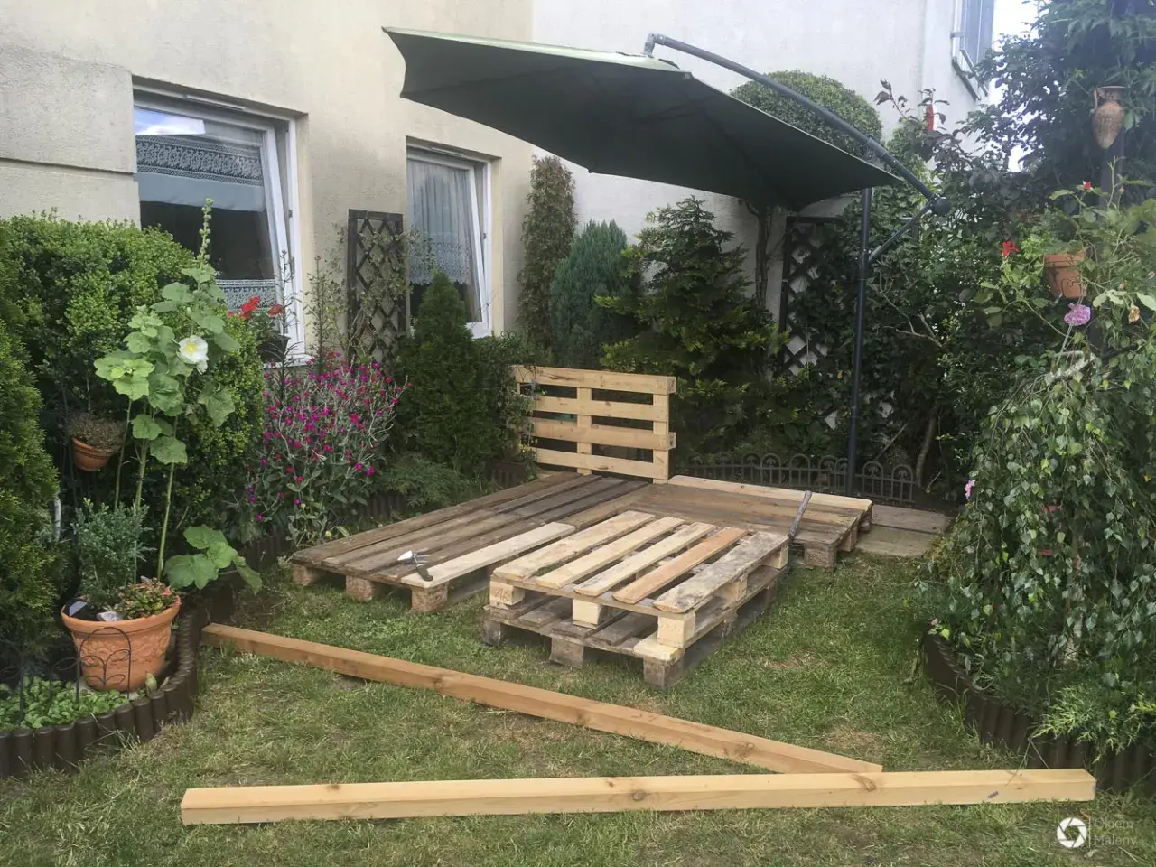 Taras z palet w trakcie budowy, otoczony zielenią i kwiatami. Pod dużym parasolem powstaje miejsce do relaksu.