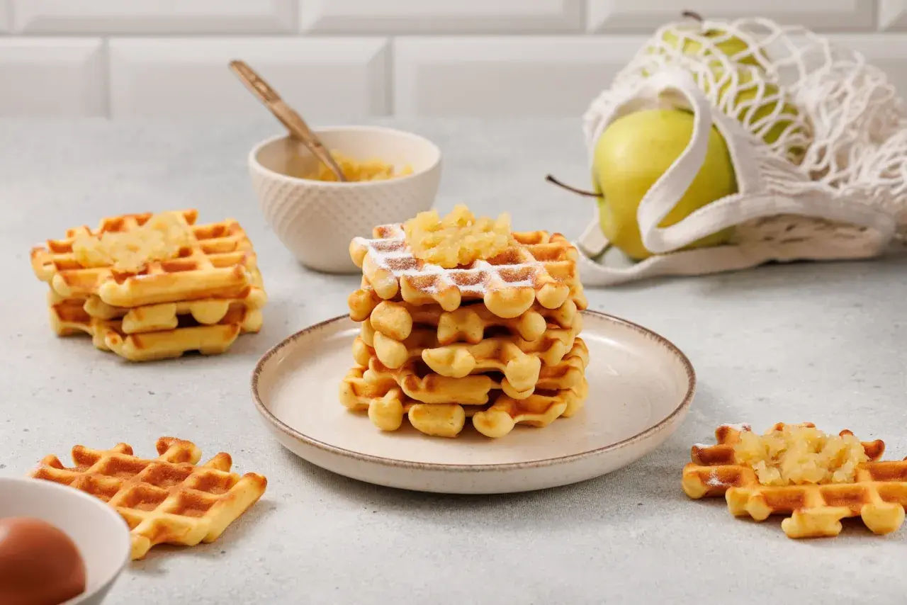 Goldbraune Waffeln mit Apfelmus bestreut, daneben eine Schüssel mit Apfelmus und ein grüner Apfel im Netzbeutel.