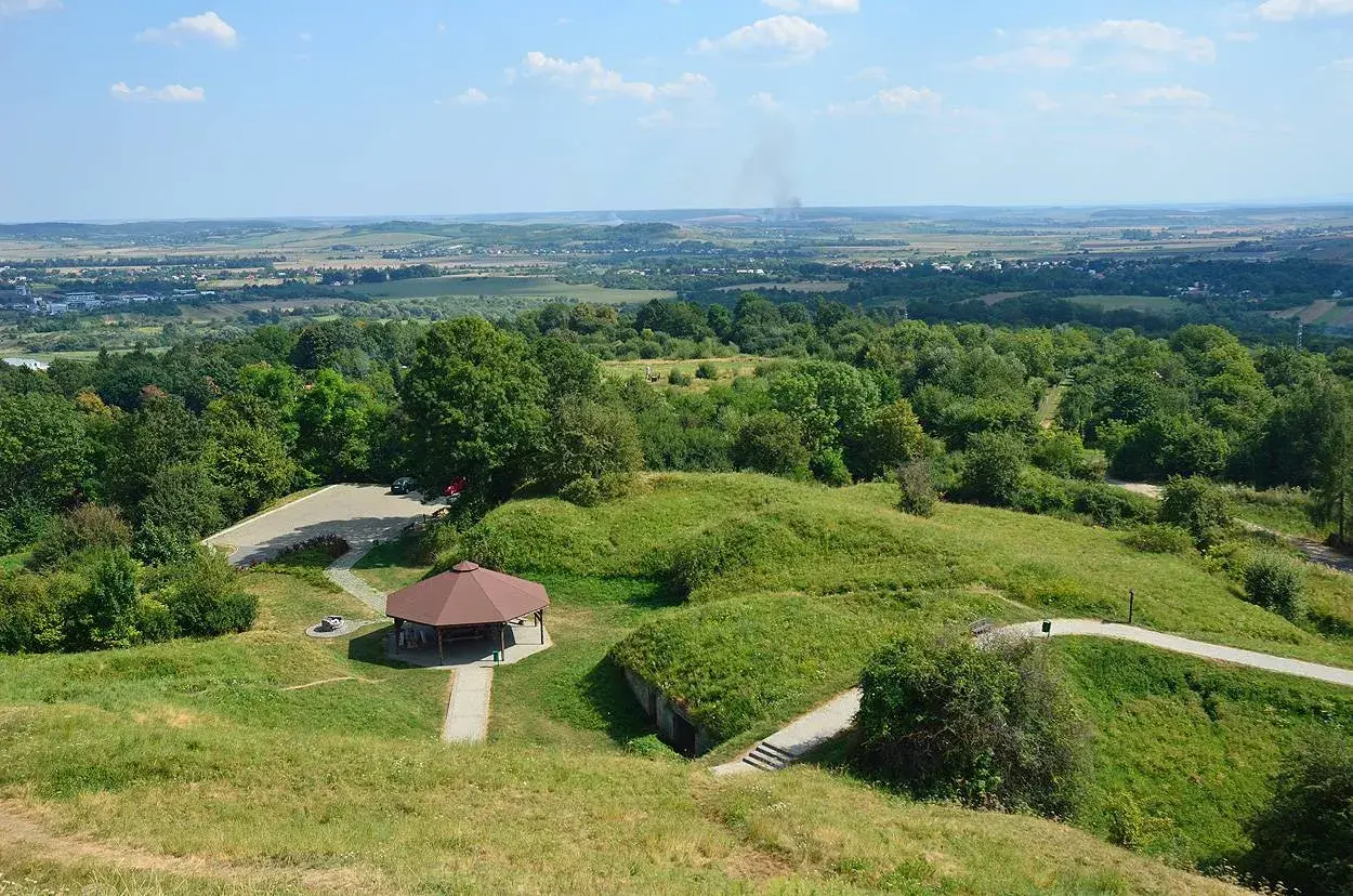 Zielony kopiec tatarski w Przemyślu z altaną i parkingiem. W oddali widać panoramę miasta i dymiące kominy.
