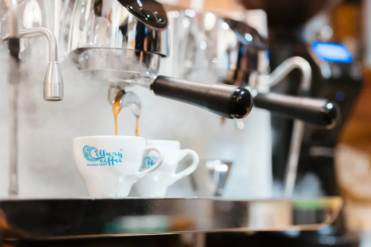 Gorąca kawa spływa do filiżanki z logo "Celberg Coffee". Zaparzacz paruje, sugerując idealne cappuccino ile ml.