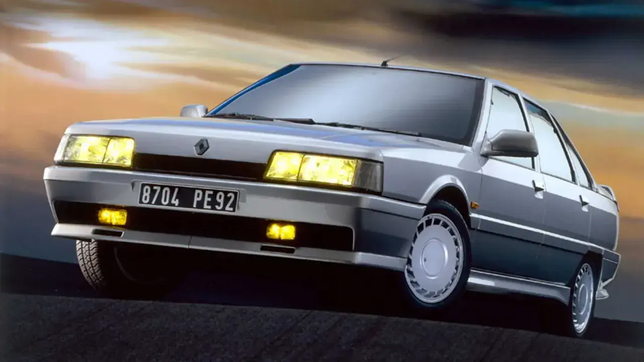 Un elegante Renault 21 Turbo plateado, con sus faros amarillos encendidos, se luce contra un cielo crepuscular.