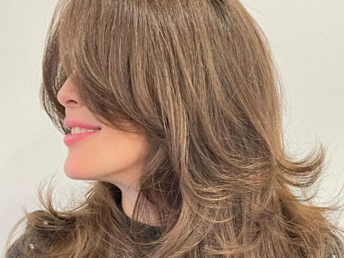 Mujer sonríe con un corte mariposa en su cabello castaño, con capas que enmarcan su rostro.
