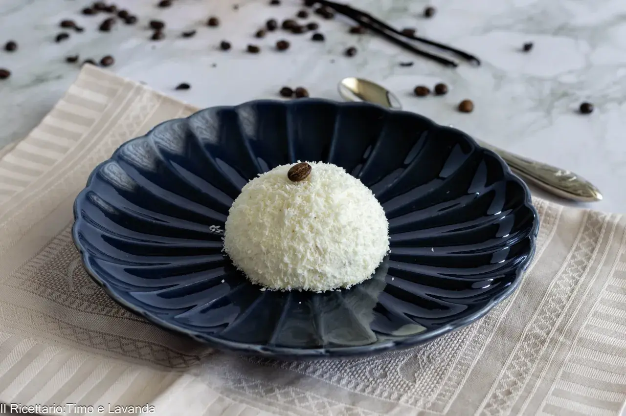 Semifreddo tartufo bianco, un dolce raffinato ricoperto di cocco e decorato con un chicco di caffè, su un piatto blu scuro.