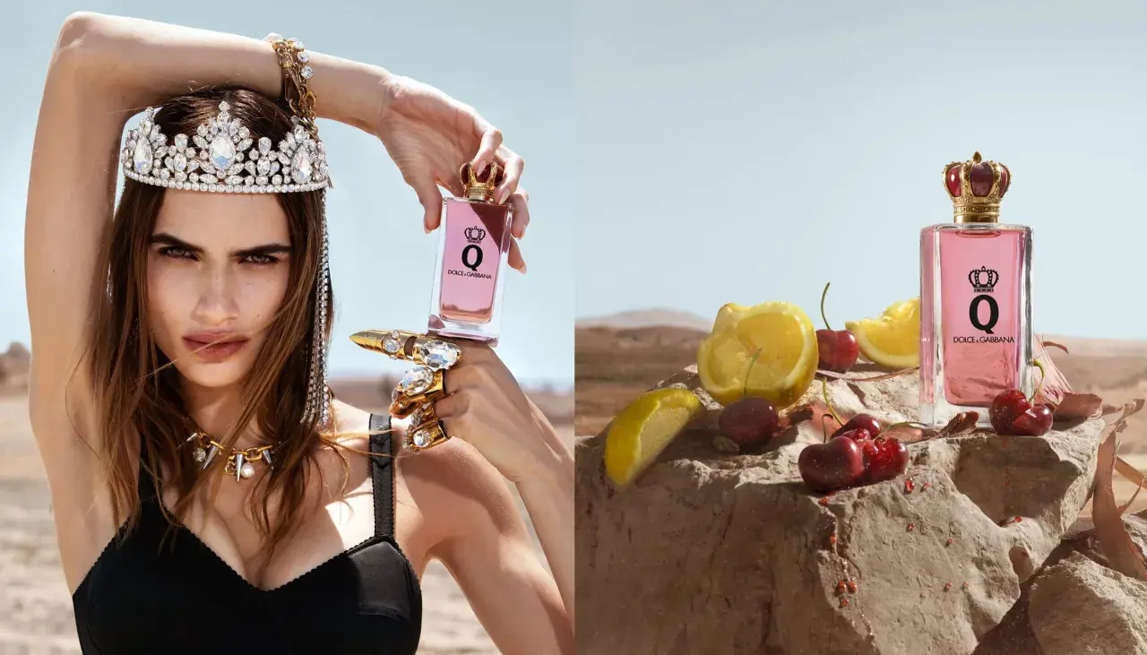 Królowa w koronie i biżuterii trzyma flakon perfum Dolce & Gabbana Q. Obok, na kamieniu, leżą wiśnie i cytryny.