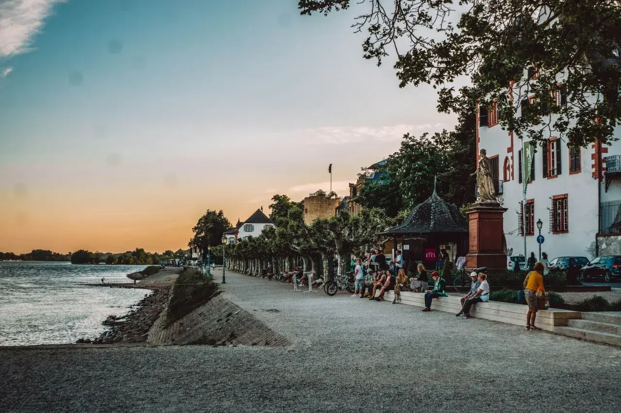 Entspannter Abend am Rhein: Menschen sitzen auf einer Promenade, ein schönes Gebäude und Bäume. Perfekt für Ausflugsziele Wiesbaden.