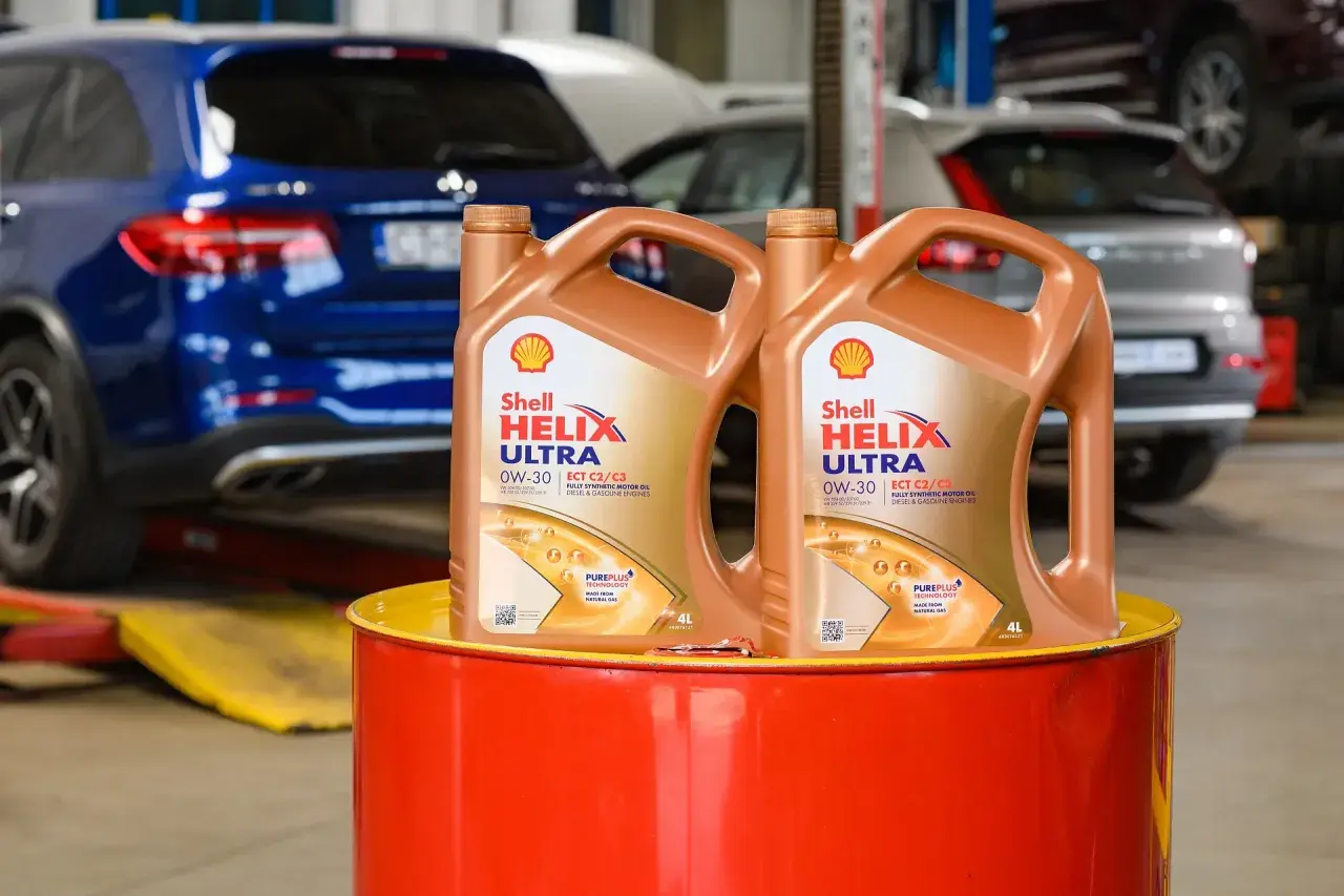 Dwa kanistry oleju silnikowego Shell Helix Ultra 0W-30 stoją na czerwonym bębnie. W tle widać samochody w warsztacie.