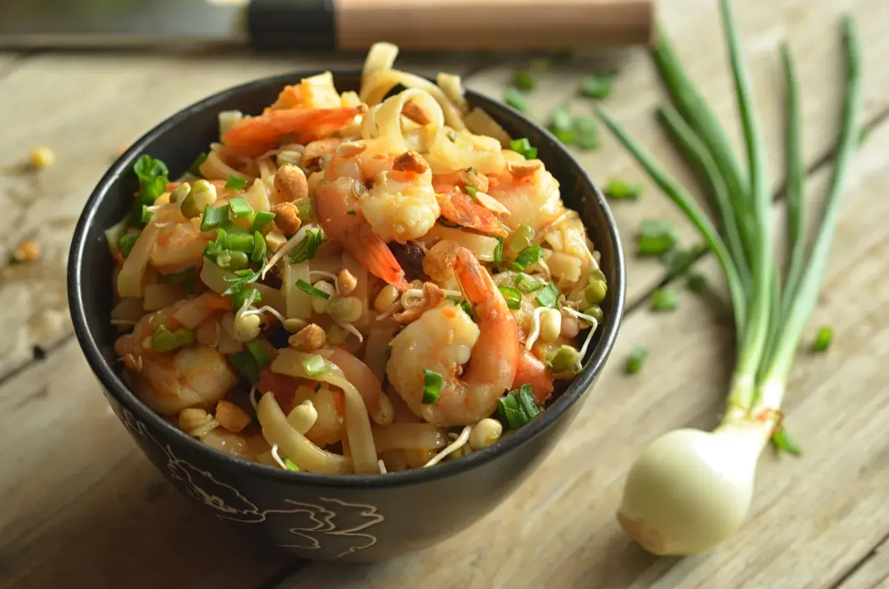 Aksamitne pad thai z krewetkami, posypane szczypiorkiem i orzeszkami, czeka w czarnej misce na drewnianym blacie. Obok leży cebulka.