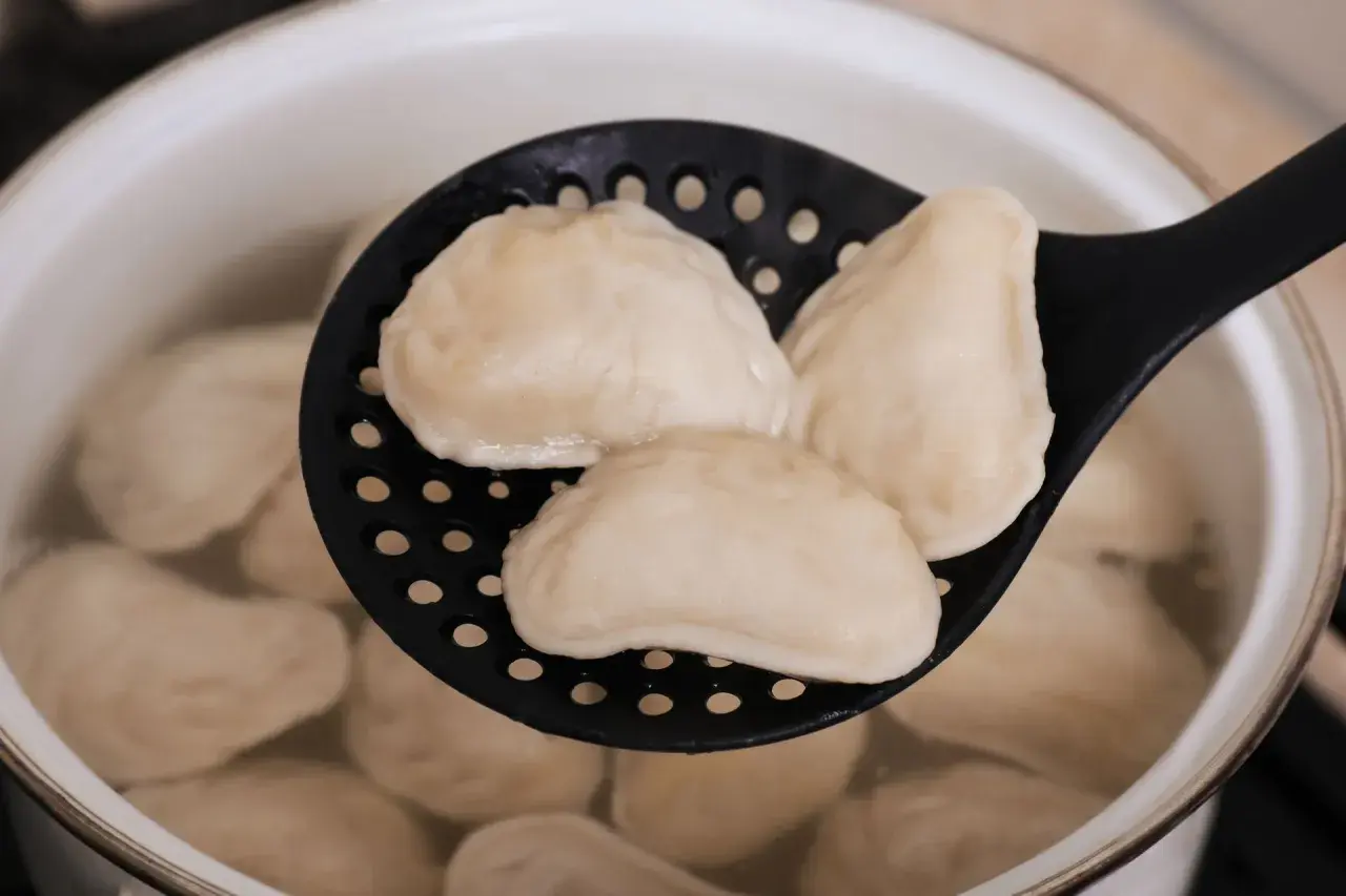 Gotowane pierogi na czarnej łyżce cedzakowej w garnku z wodą.