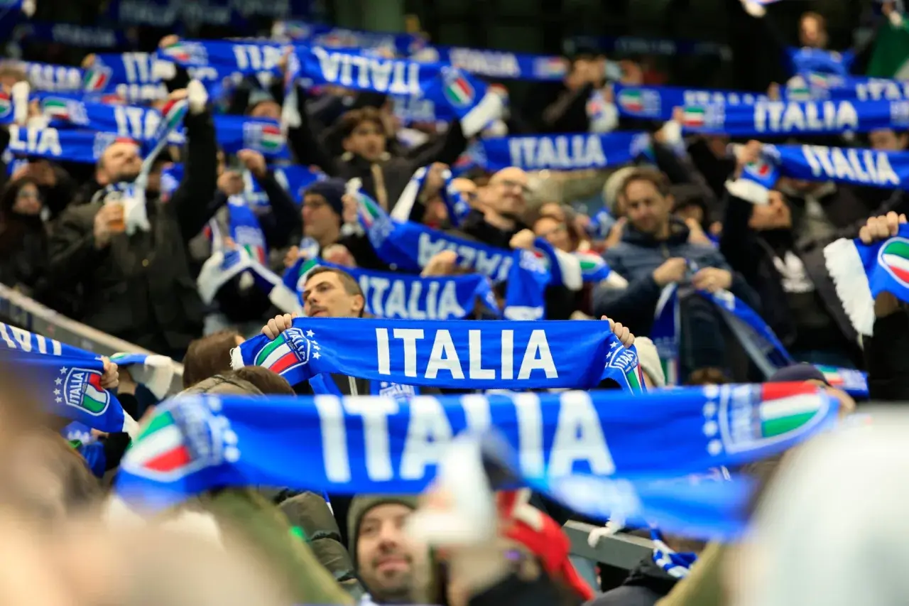 Tłum kibiców na meczu, trzymający niebieskie szaliki z napisem "ITALIA" i flagą Włoch. Emocje towarzyszące kibicowaniu podczas mistrzostw świata.