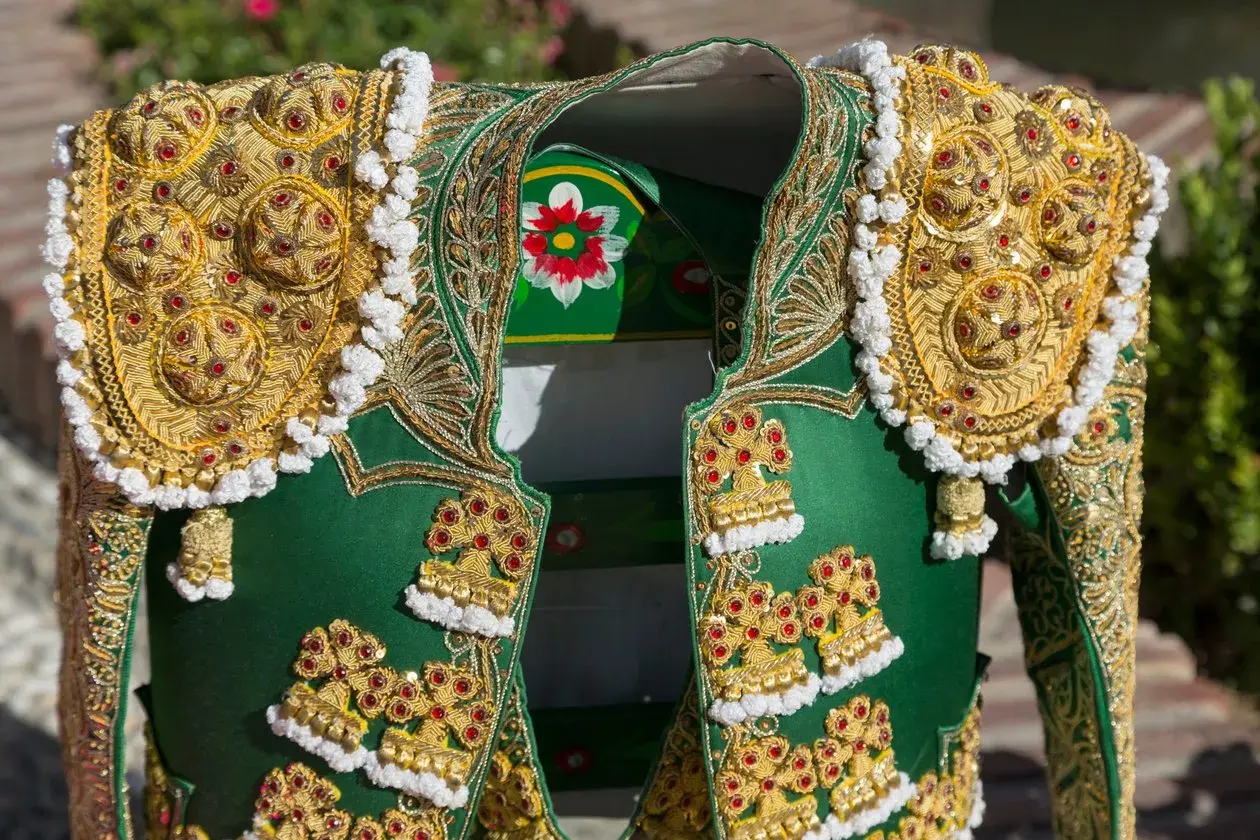Detalle de un traje de luces de torero, con bordados dorados y pedrería roja sobre tela verde.