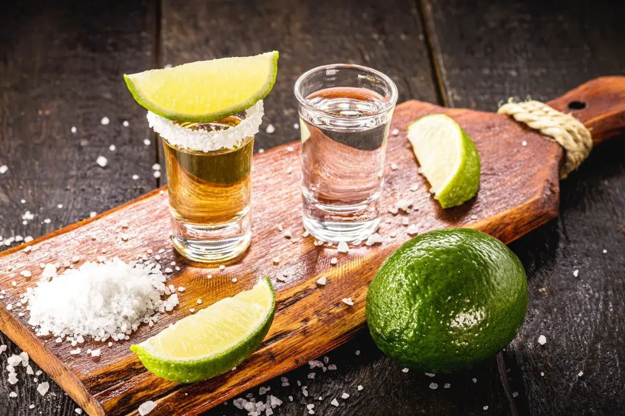 Zwei Tequila-Gläser, Salz und Limettenspalten auf einem Holzbrett. Die Tequila-Reihenfolge: zuerst Salz, dann Tequila, dann Limette.