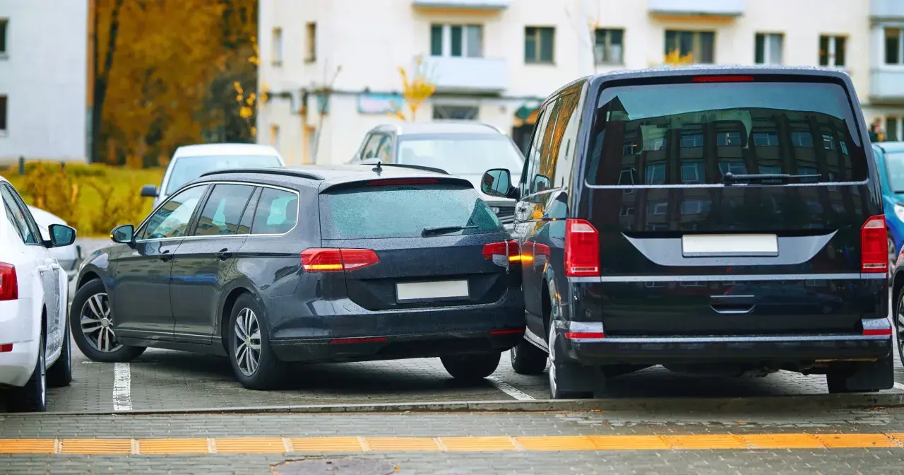 Ciemne kombi i van stoją blisko siebie na parkingu. Widać fragment białego auta. To strefa ruchu, gdzie parkowanie wymaga uwagi.