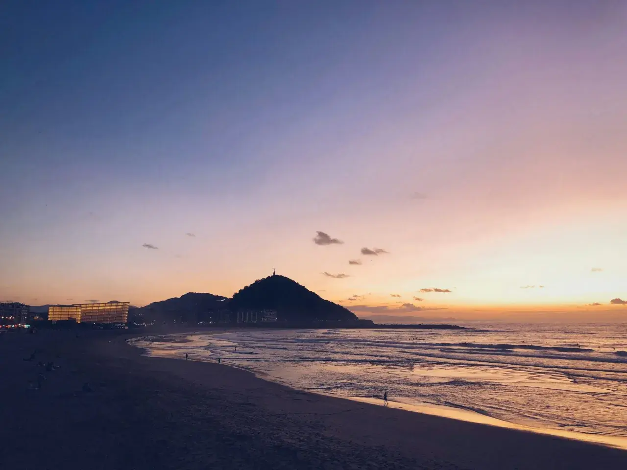 Atardecer en la costa San Sebastián. El cielo se tiñe de tonos rosados y naranjas sobre el mar y la playa, con la silueta del monte Urgull al fondo.