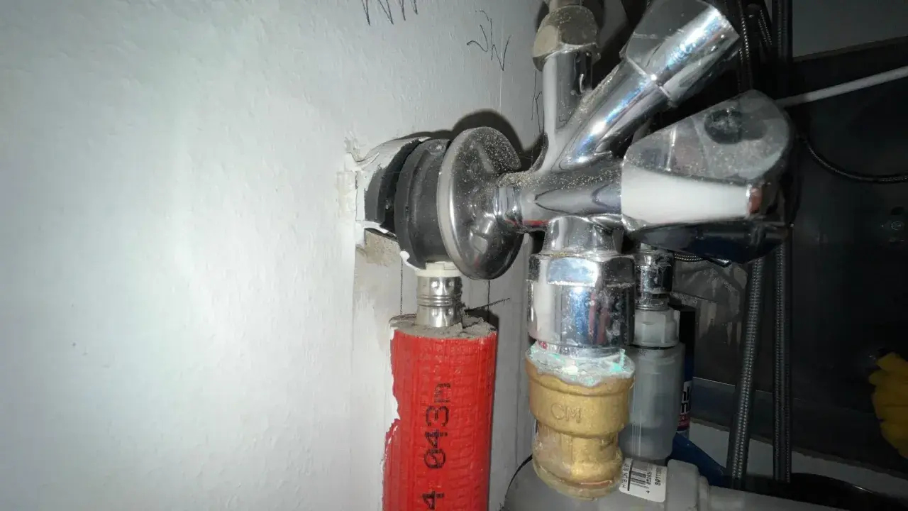 Chromglänzendes Eckventil der Wasserleitung mit rotem Schutzschlauch und sichtbaren Leitungen an einer weißen Wand.