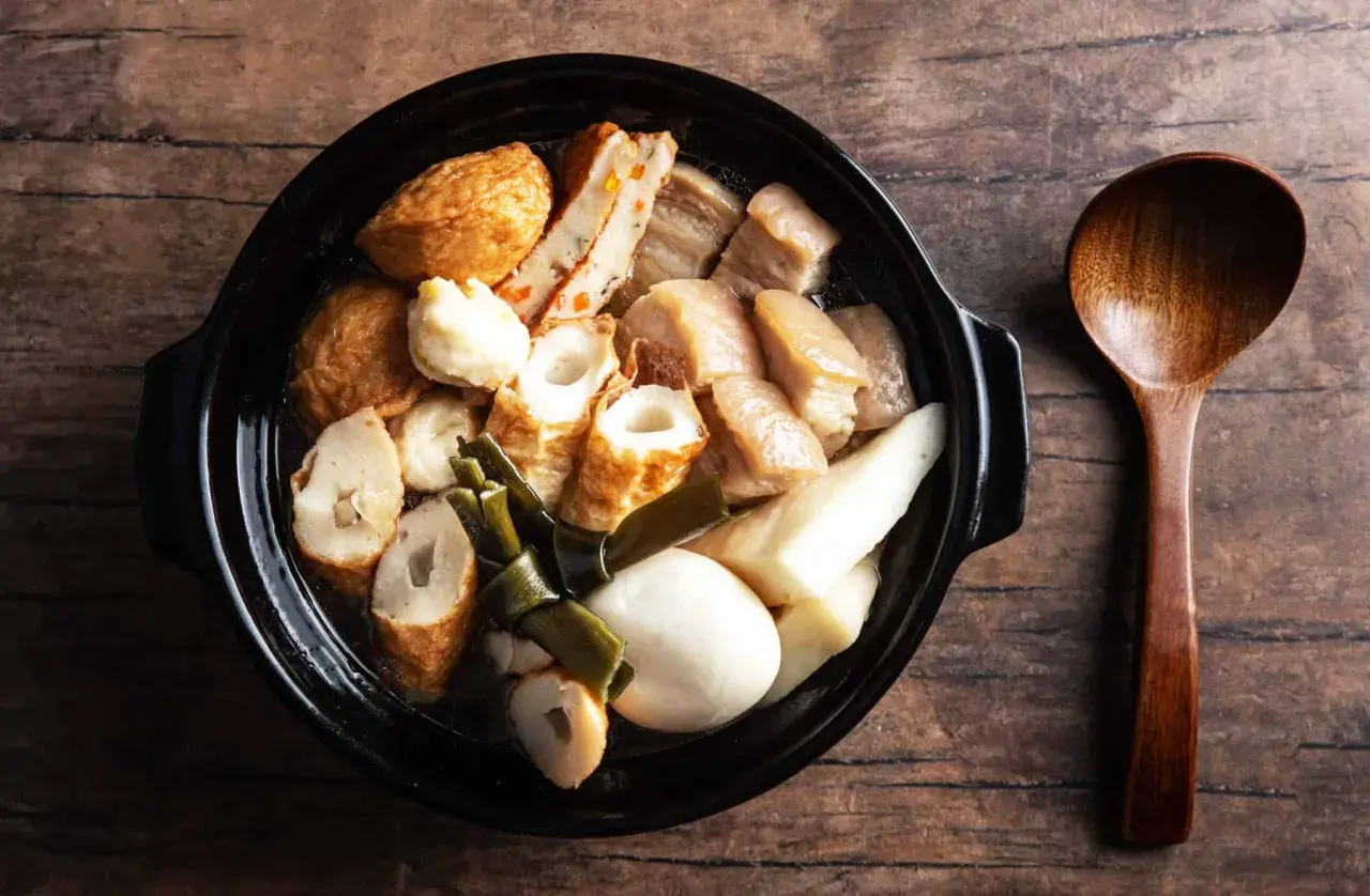 A steaming pot of oden, a Japanese fish cake stew, with various ingredients like daikon and pork belly, served with a wooden spoon.