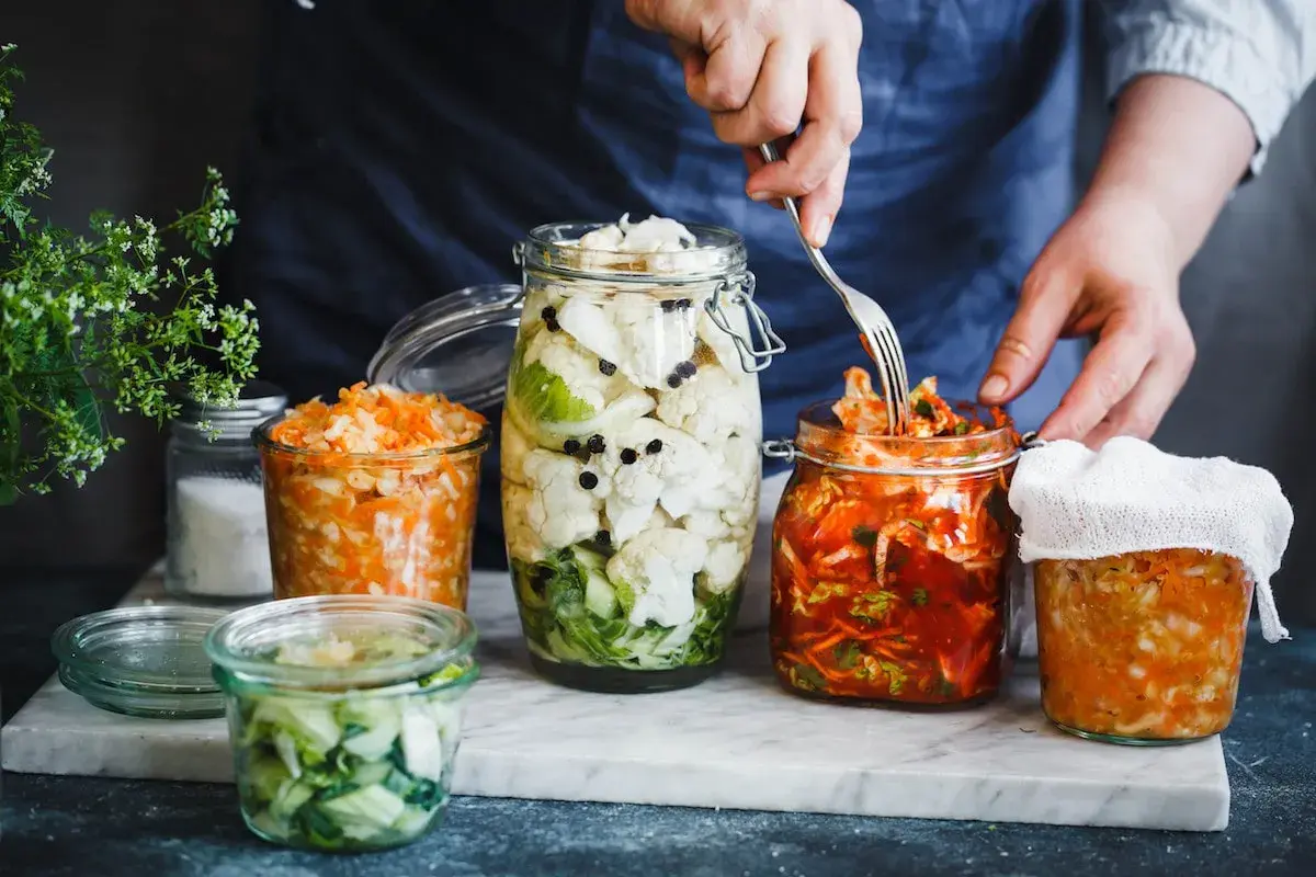 Kimchi selber machen im Glas: Verschiedene fermentierte Gemüse in Gläsern, darunter ein Glas mit rotem Kimchi, das gerade mit einer Gabel entnommen wird.