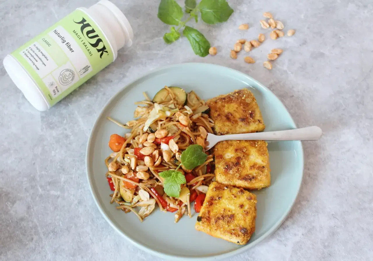 Tallrik med nudlar, grönsaker och friterad tofu. Bredvid en burk HUSK Naturlig fiber, perfekt för mat utan gluten och en balanserad mage.