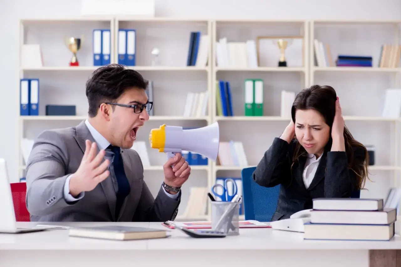 Uomo urla con megafono contro collega che si tappa le orecchie. Un chiaro esempio di **test stress da lavoro**.