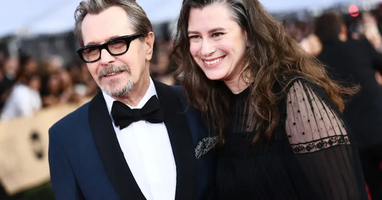 Gary Oldman z żoną, oboje uśmiechnięci, on w eleganckim garniturze, ona w czarnej sukni z koronkowymi rękawami.