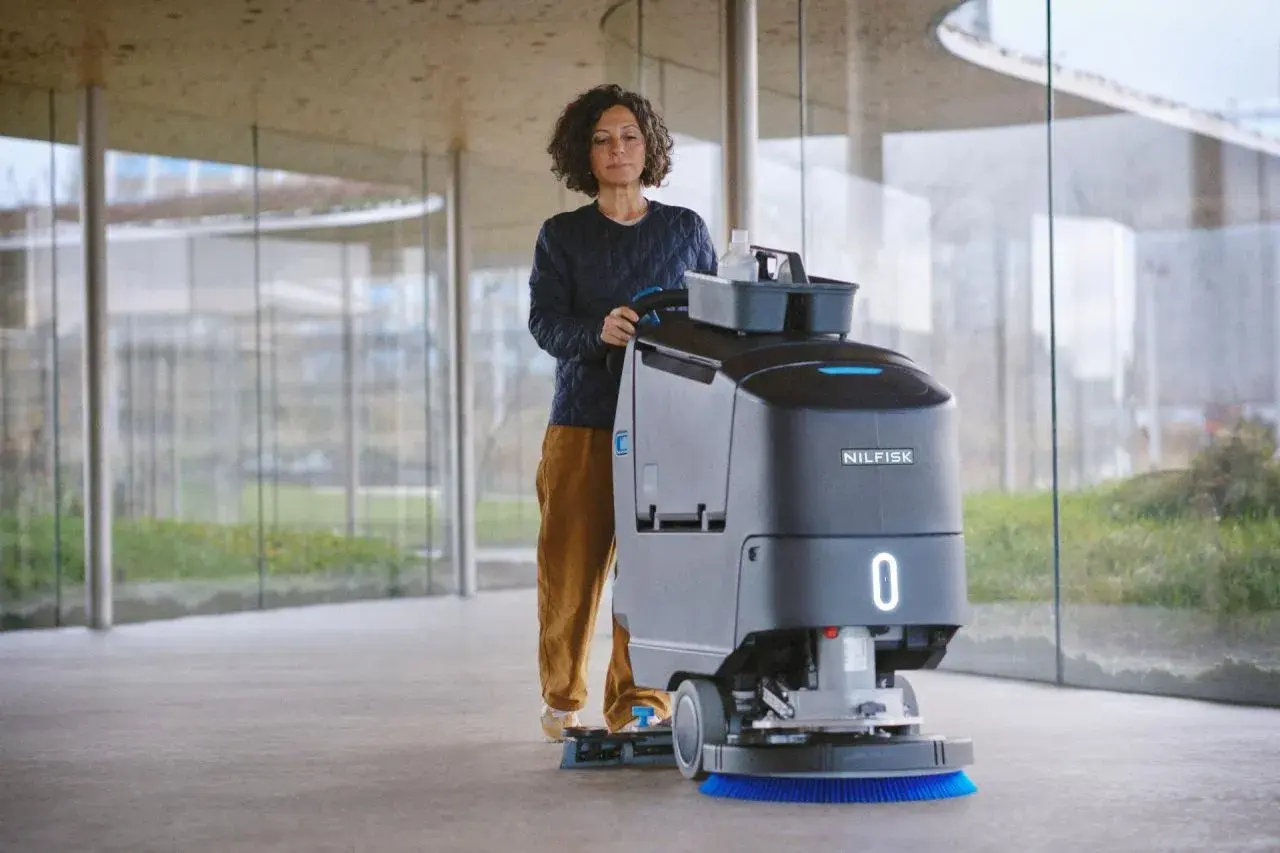 Mujer operando una máquina de Nilfisk, parte de sus avanzados sistemas de limpieza, en un pasillo moderno con grandes ventanales.