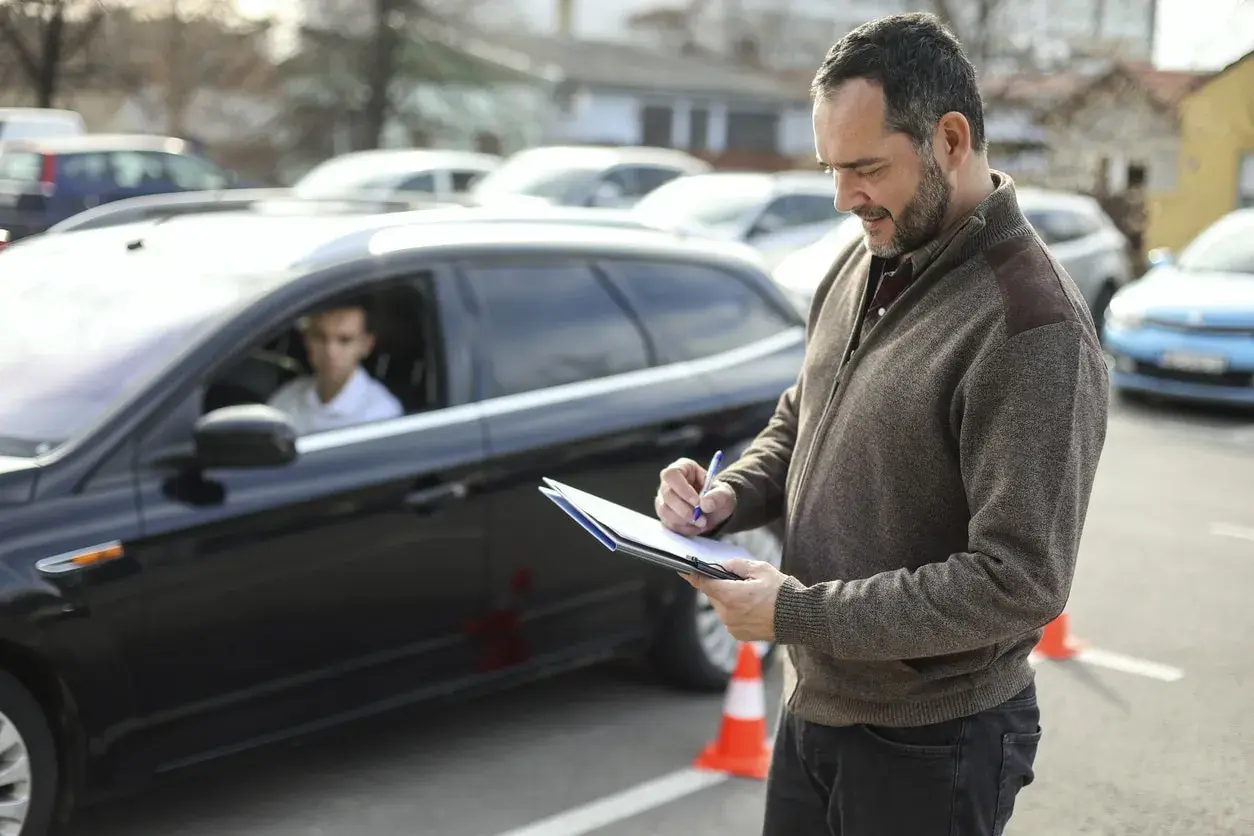 Examinador anotando en una carpeta. Un joven conductor espera en un coche negro. El examen práctico DGT y su precio son importantes para obtener el carnet.
