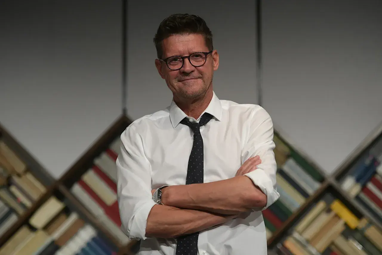 Wojciech Malajkat w białej koszuli i krawacie, z okularami na nosie, stoi na tle regałów z książkami. Jego filmy to klasyka polskiego kina.